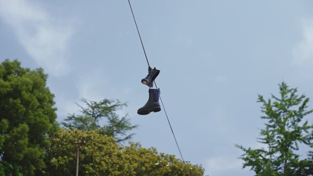 A pair of black boots dangling from an overhead wire against a soft, blue sky and leafy green trees, capturing an enigmatic and urban-rustic juxtaposition that hints at stories left behind.