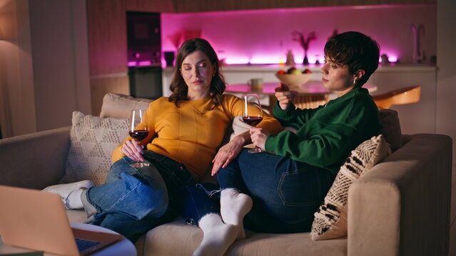 Two girls relaxing sofa holding glasses red wine discussing laptop movie closeup