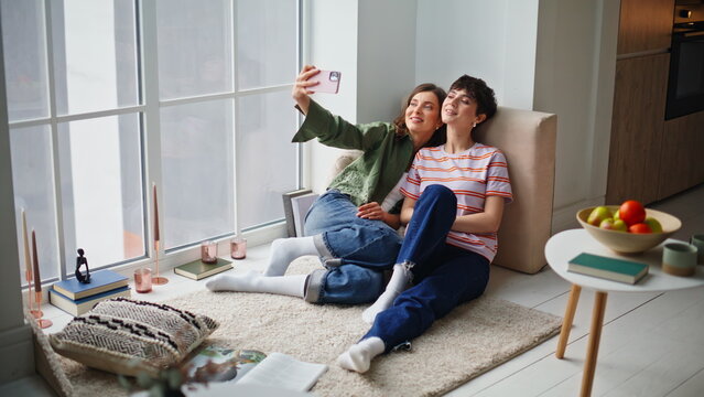 Girls taking cellphone picture sitting apartment floor. Smiling two women 