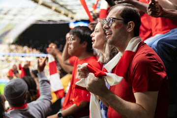 Obraz premium Anxious football supporters watching a decisive final attack during match from packed stadium stands with red scarves and fan tension. Supporter holding breath with nervous anticipation.