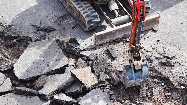 Orange mini excavator breaking asphalt road surface at urban construction site