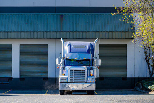 Blue classic big rig semi truck tractor with chrome parts standing on the warehouse dock loading cargo for the next route