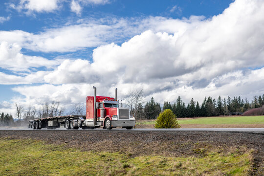 Classic red big rig semi truck tractor with extended cab and empty flat bed semi trailer running on the narrow road with water dust after the rain