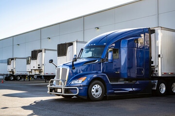 Naklejka na ściany i meble Dark blue big rig semi truck with grille guard loading cargo into refrigerated semi trailer standing in the warehouse dock gate