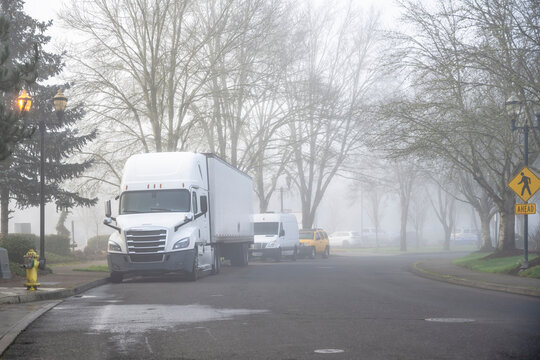 High cab white big rig semi truck with dry van semi trailer standing on the city street with foggy weather waiting for the next freight