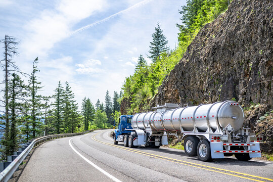 Day cab blue big rig semi truck tractor transporting liquid cargo in long armored tank semi trailer running on the winding mountain road with rock cliff on the side