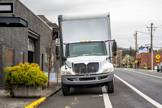 Middle duty white rig semi truck with bo trailer standing on the city street road making cargo delivery to the business sides