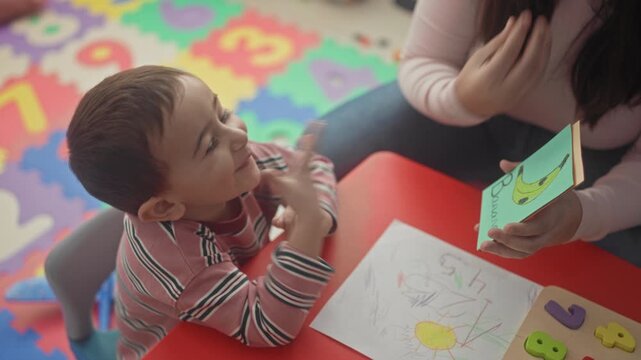 Woman holds orange card and points to child's drawing on paper at a small red table in a building; early learning curiosity.