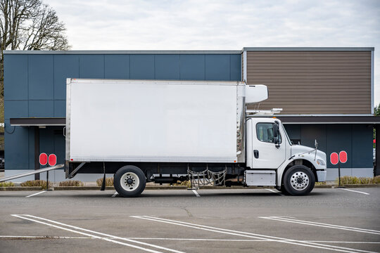 Profile of the day cab middle duty rig semi truck with refrigerated box trailer making delivery standing on the parking lot in front of the business building