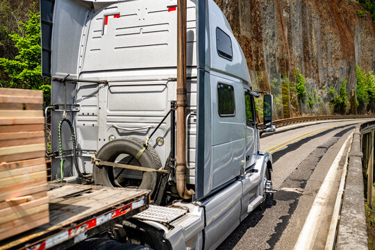 White powerful big rig semi truck tractor with extended cab transporting fastened wood lumber on flat bed semi trailer running on the narrow mountain road