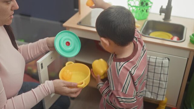 Boy holding yellow toy pot and green lid while reaching into toy kitchen cabinet in playroom with woman assisting; nurturing bonding.