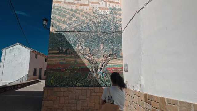 Woman Admires a Large Mural of Religious Art and Olive Trees in a Traditional Spanish Village