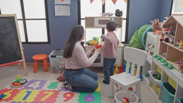 Woman kneeling holds plastic stacking cups and hands a cup to a little boy while arranging a play kitchen in a childcare building; playful bonding.