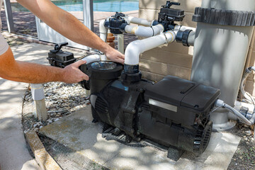 Close-up horizontal view of a technician's hands installing or servicing a black variable speed pool pump motor connected to PVC plumbing pipes and a filtration system on an outdoor concrete pad, USA © Andrei Liashchou