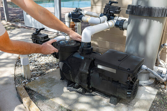 Close-up horizontal view of a technician's hands installing or servicing a black variable speed pool pump motor connected to PVC plumbing pipes and a filtration system on an outdoor concrete pad, USA