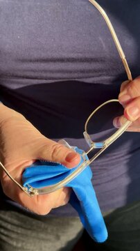 Closeup hands cleaning eyeglasses with satin cloth, woman gently polishes gold frames and lenses, sunlight highlights microfiber