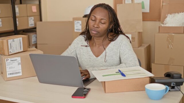 Woman typing on laptop and checking order form among stacked shipping packages and cup on desk in a building; entrepreneurial focus.