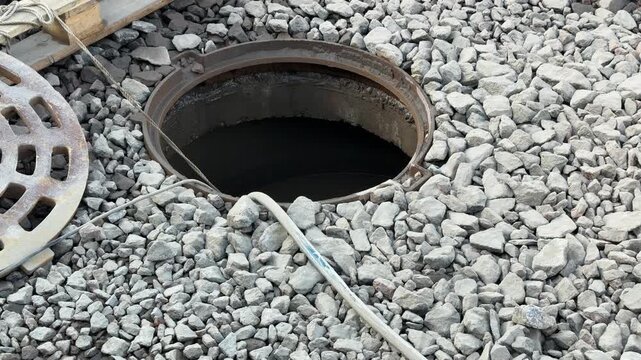 Open inspection well with rubber hose, gravel street, wooden pallet and shopping cart, pump engine running as hose drains water
