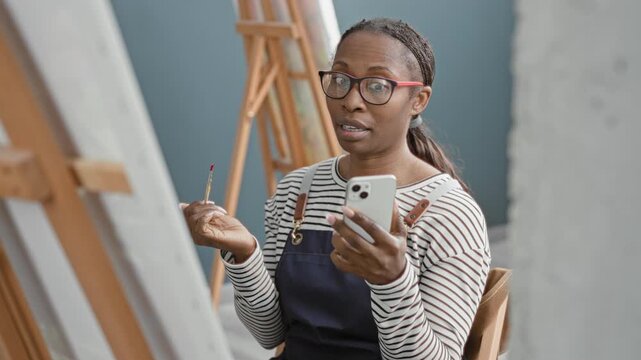 Woman in apron holds smartphone and paintbrush, gestures to canvas and speaks while seated at an easel in a studio; creative teaching.