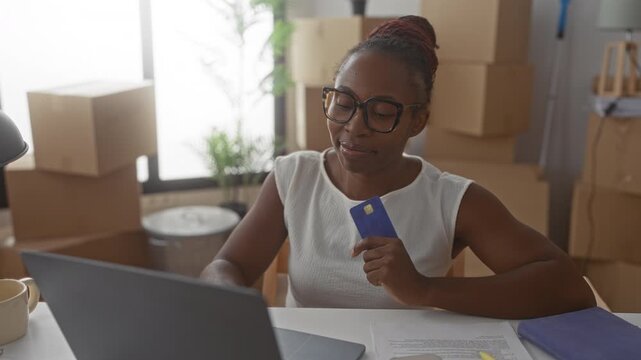 Young woman holding blue credit card smiles at laptop amid stacked moving boxes inside building; optimism new start.