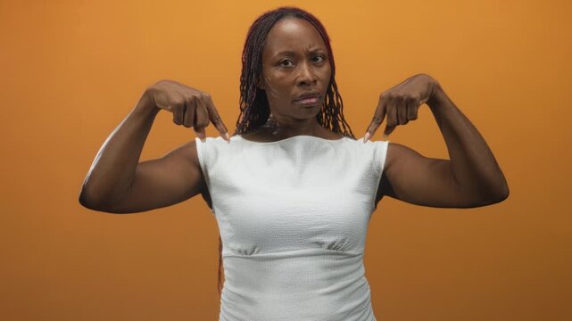Young african american woman in white dress pointing both fingers at her upper arms and chest in studio with orange backdrop; confidence.