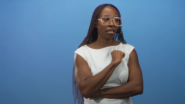 Young african american woman bites knuckle and crosses arms in studio wearing white sleeveless dress and glasses; concern reflection.