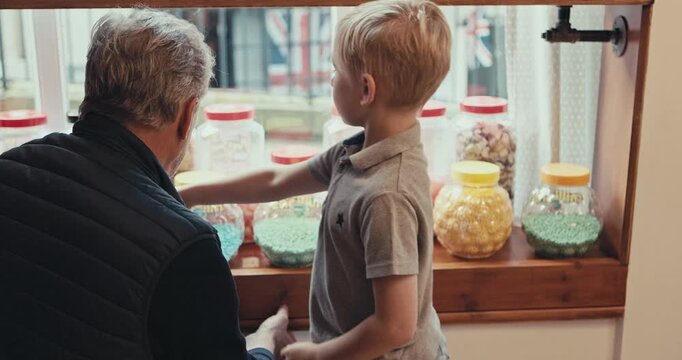 Grandfather, shopping and choice of candy for a child at a shop or sweets store shelf. Senior man and a boy kid or customer talking and buying sugar treats in window display jar on travel trip