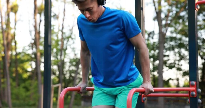 Athletic young man with headphones exercising on parallel bars at outdoor gym