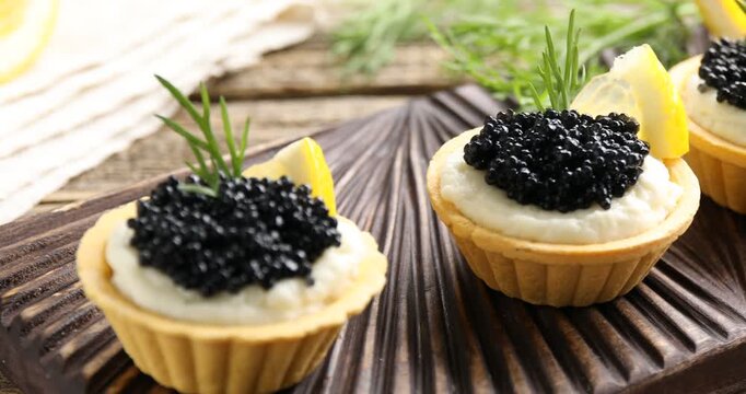 Tasty tartlets with black caviar, cream cheese, dill and lemon on wooden table, closeup. Camera moving right