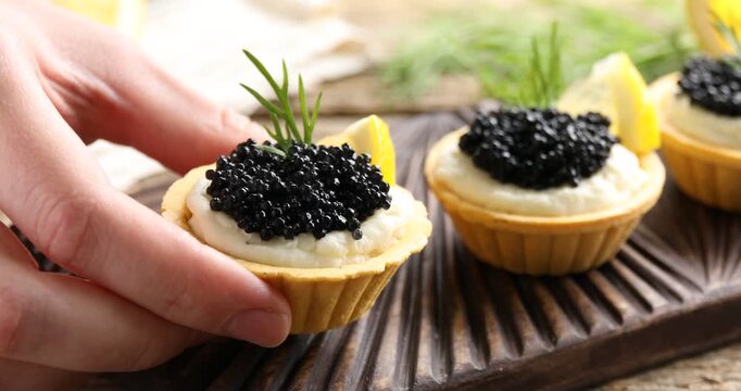 Woman taking tasty tartlet with black caviar and cream cheese at wooden table, closeup