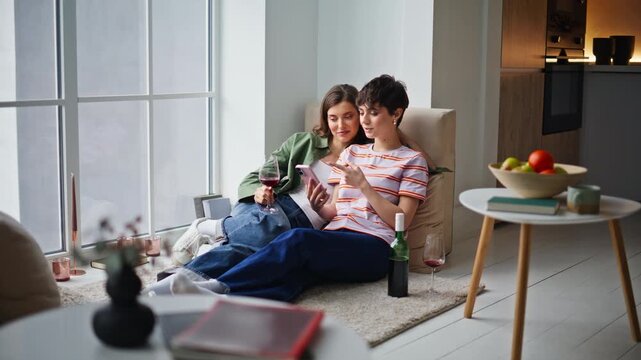 Bonding women watching cellphone sitting cozy apartment floor with wine glasses.