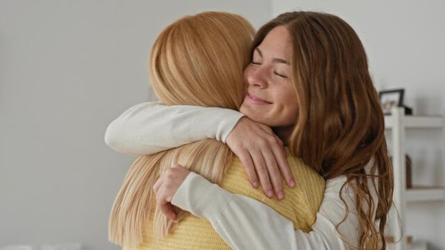 Woman hugging another woman with hands on shoulder and arms around back in a home living room with shelf and plant, yellow sweater visible; comfort reunion.