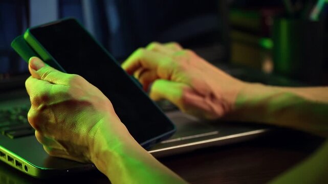 Business woman holding mobile phone in her hands sitting at work table with laptop. Online work, study, research, connection on pc. Closeup workspace in creative art green yellow dark shadow light