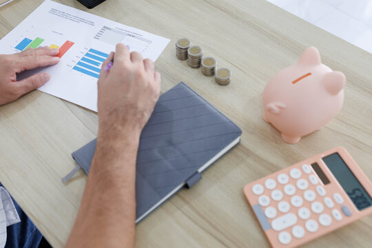 
Man sits at desk, thoughtful, pen in hand. Piggy bank and coins rest nearby, symbolizing savings. He sketches ideas on notebook, surrounded by charts. Office shelves hold colorful folders