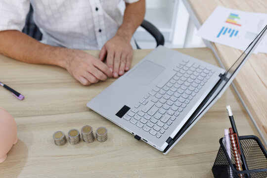 
Man sits at desk, thoughtful, pen in hand. Piggy bank and coins rest nearby, symbolizing savings. He sketches ideas on notebook, surrounded by charts. Office shelves hold colorful folders