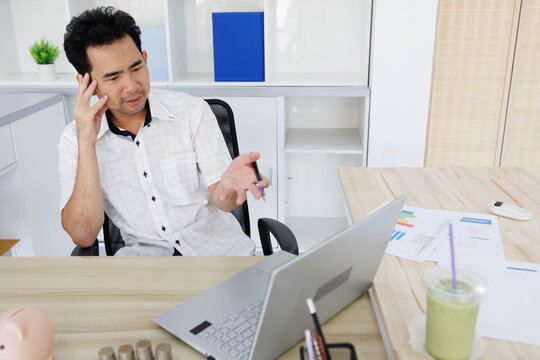 
Man sits at desk, thoughtful, pen in hand. Piggy bank and coins rest nearby, symbolizing savings. He sketches ideas on notebook, surrounded by charts. Office shelves hold colorful folders