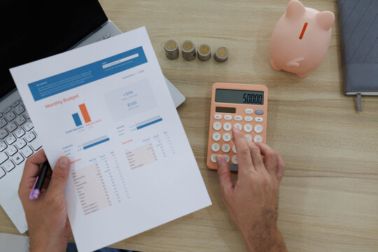
Man sits at desk, thoughtful, pen in hand. Piggy bank and coins rest nearby, symbolizing savings. He sketches ideas on notebook, surrounded by charts. Office shelves hold colorful folders