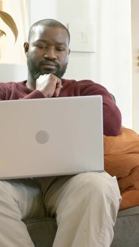 Vertical video: Reading laptop content, maroon-clad man tapping trackpad on sofa working smartwatch