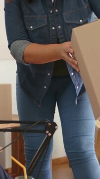 Vertical video: Entering woman in denim carrying box at desk, avoiding lamp, placing box to unpack