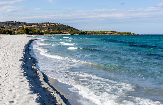Beach at S'Ena e Sa Chitta in Capo Comino, Sardinia, Italy. Sandy shoreline with turquoise waves and scenic coast. Sunny day with clear blue sky and green hills in the background.