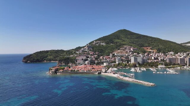 Budva Old Town rises from Adriatic coast with marina, red roofed buildings and stone pier set against turquoise water and forested hills, captured from drone for panoramic coastal city view
