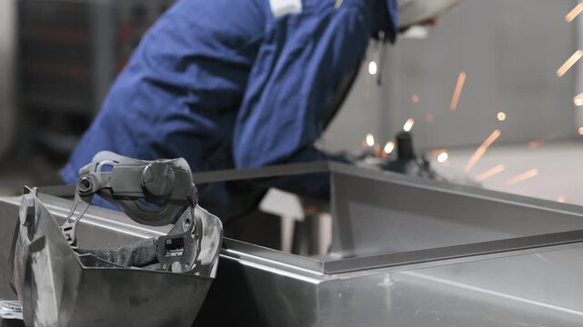 Industrial worker grinding metal frame with angle grinder in factory workshop. Sparks flying during steel processing and metal fabrication.