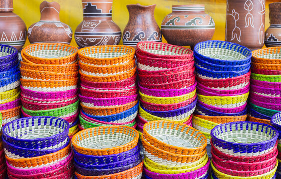 Cestos de mimbre tejidos a mano en m&uacute;ltiples colores brillantes, apilados verticalmente en exhibici&oacute;n, atr&aacute;s jarrones de barro con grecas prehisp&aacute;nicas 