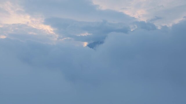Soft blue sky transitions through varying cloud densities as wispy white formations drift gently across the frame, revealing glimpses of a distant mountain peak shrouded in atmospheric haze and mist.