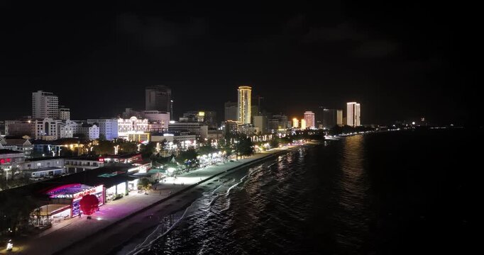 Forward aerial night view of Sihanoukville shows a vibrant coastline, where illuminated high-rise hotels and beachside businesses line the sandy shore as waves reflect the glowing cityscape.
