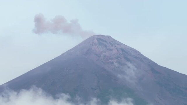 Guatemala active Fuego volcano releases volcanic smoke and ash from its crater against pale blue sky. White clouds drift along rugged mountain slopes as steam rises from the peak.