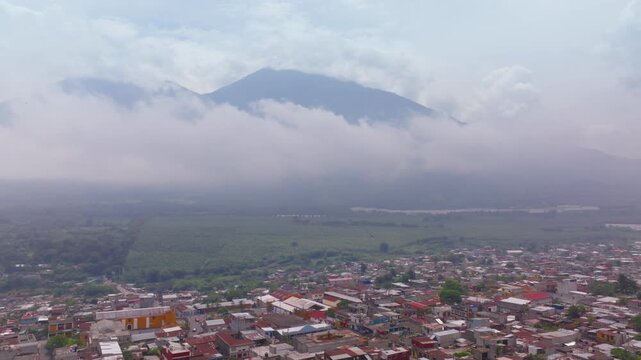 Aerial drone footage captures a scenic Guatemalan town with colorful rooftops nestled in a valley. Misty clouds drift around a majestic volcano peak while lush green vegetation surrounds urban area.