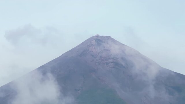 Majestic Fuego volcano in Guatemala emits volcanic ash and smoke from its rugged summit while wispy clouds drift across the dark rocky slopes under a soft pale blue overcast sky during daytime.