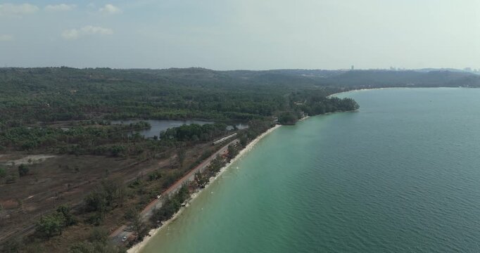 Forward aerial view of a train traveling on tracks beside a coastal road, separated by trees from the turquoise sea along a scenic shoreline.