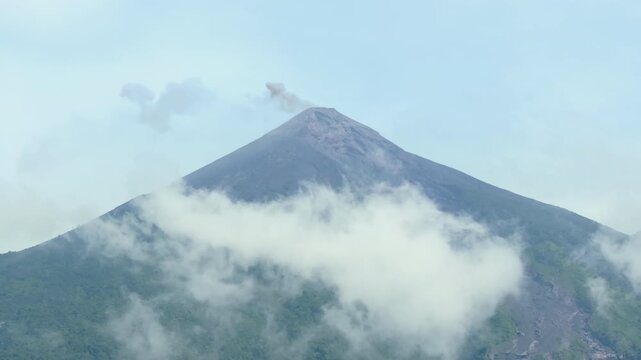 Majestic active Fuego Volcano in Guatemala releases volcanic ash and smoke from its conical peak while wispy white clouds drift across the lush green forested slopes under a pale blue hazy sky.